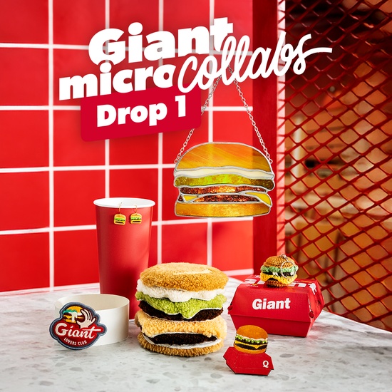Six premières créations colorées, disposées sur une table blanche dans un restaurant Quick : patch fixé au carton blanc classique qui entoure le Giant, boucles d’oreilles accrochées à un gobelet rouge, magnet, set de 7 sous-verres, vitrail avec chaîne à m